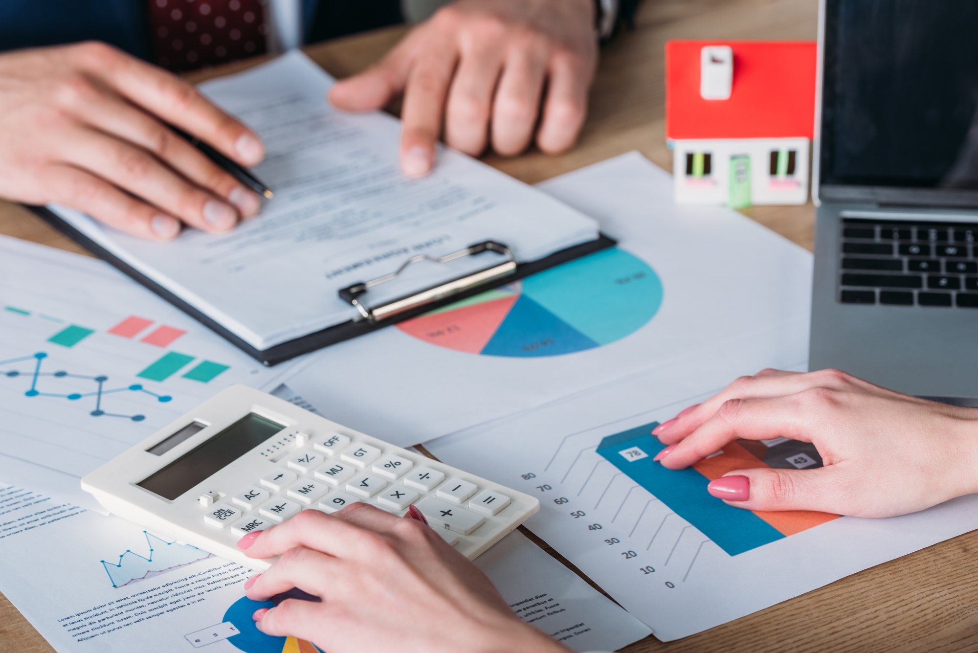 Partial view of man studying loan agreement and businesswoman using calculator while sitting at workplace with house model, graphs and charts