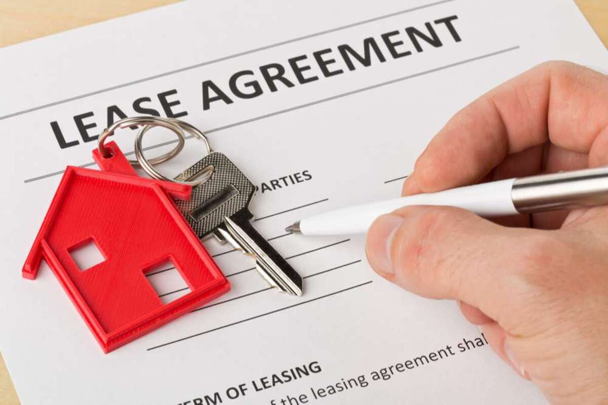 Man holding pen over house door key with red keychain pendant and lease agreement form on wooden desk